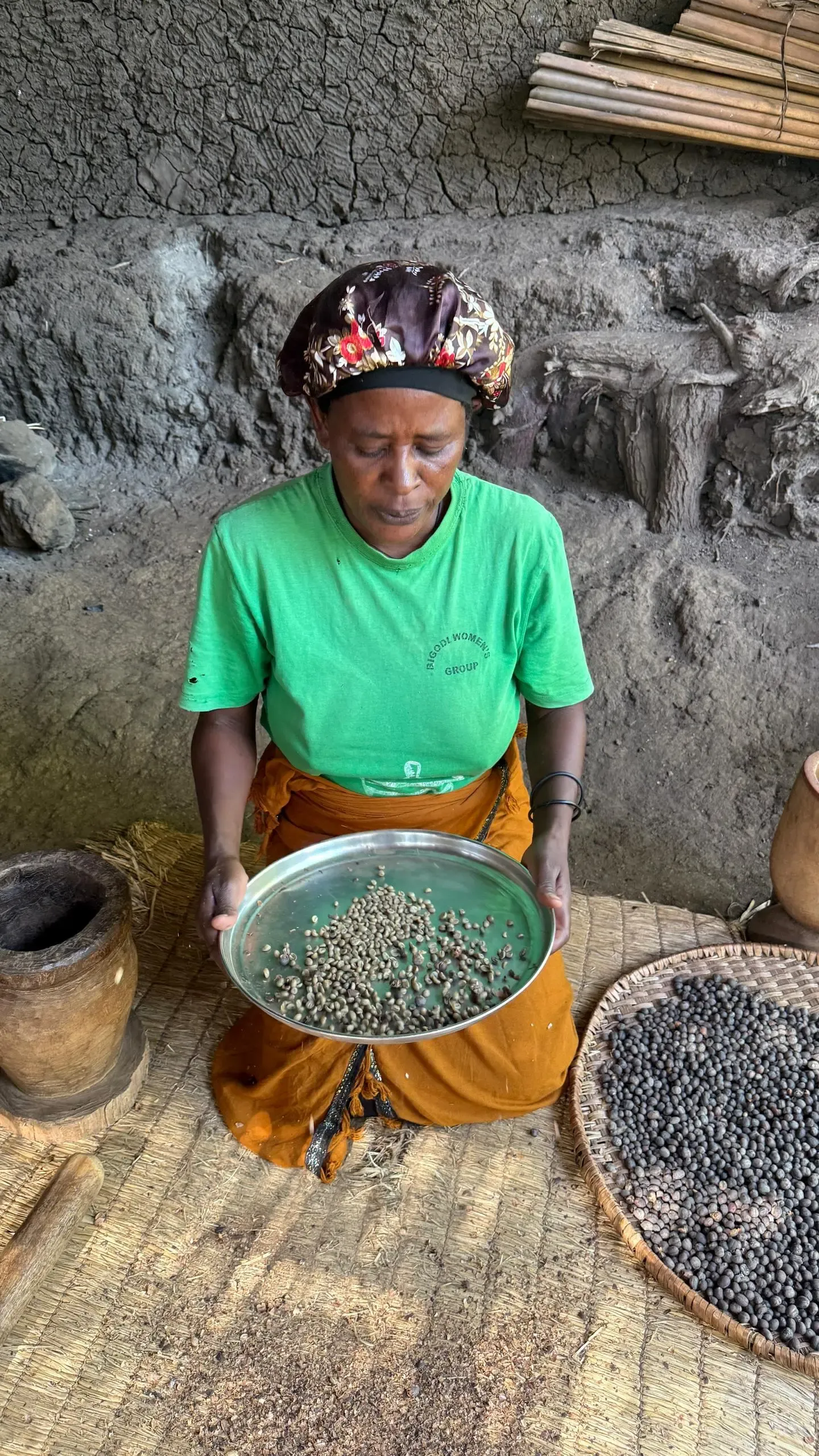 A member of the Bigodi Women's Group kneeling on a mat, holding out a metal tray of green coffee beans, a basket of dried coffee cherries beside her.