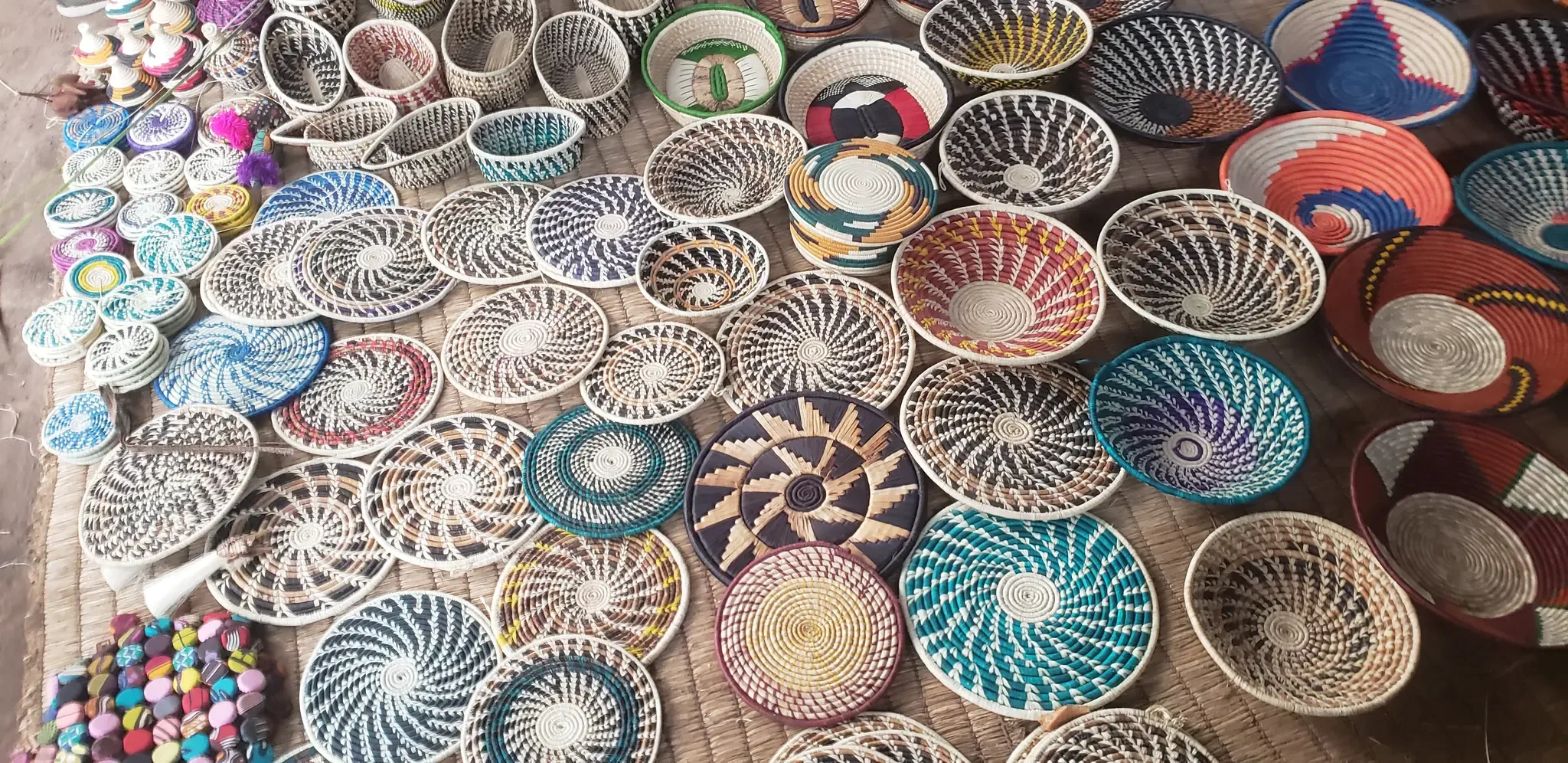 Dozens of coiled baskets and woven plates laid out on a mat, in many colours and spiral patterns.