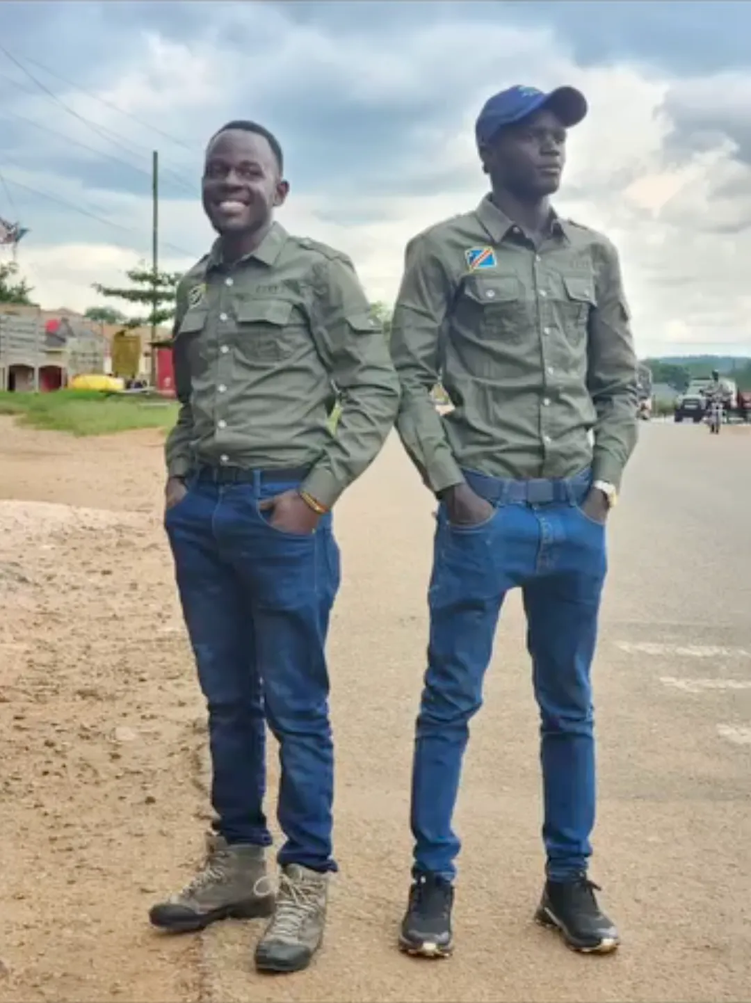 Two Bigodi guides in olive field shirts standing at the roadside, hands in pockets, under a bright clouded sky.